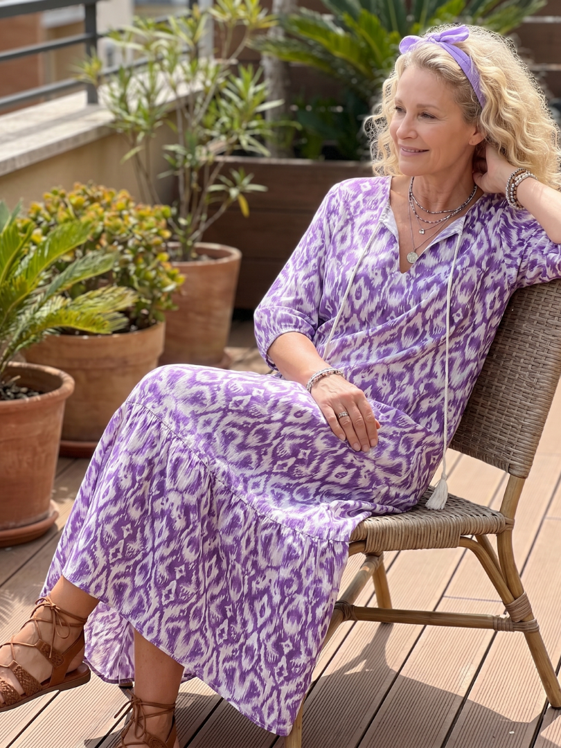 Penelope printed midi dress in purple, worn seated on a plant-filled terrace