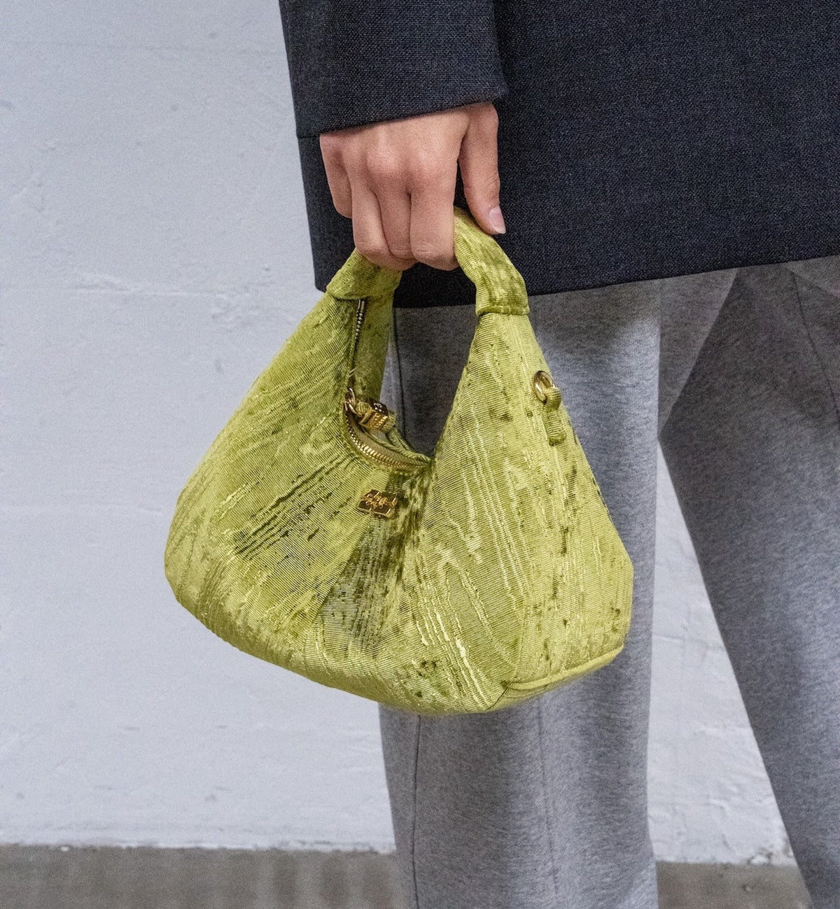 Roma Luxe green mini bag carried by the top handle beside grey trousers